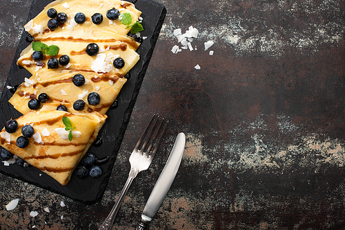top view of tasty crepes with blueberries, mint and coconut flakes served on board near cutlery on textured background
