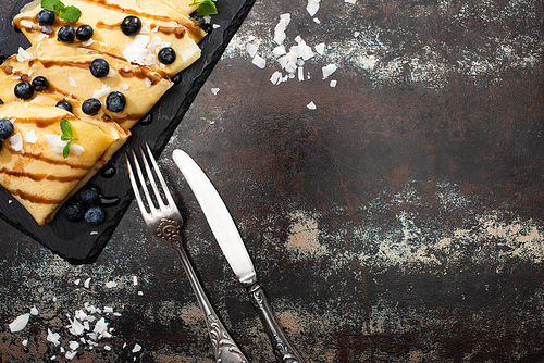 top view of tasty crepes with blueberries, mint and coconut flakes served on board near cutlery on textured background