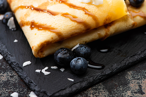 close up view of tasty crepes with blueberries and coconut flakes served on board on textured background