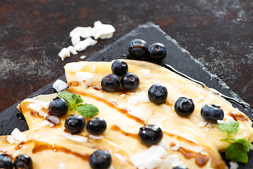 close up view of tasty crepes with blueberries, mint and coconut flakes served on board on textured background