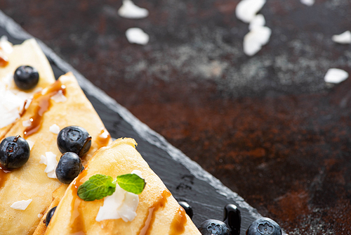close up view of tasty crepes with blueberries, mint and coconut flakes served on board on textured background