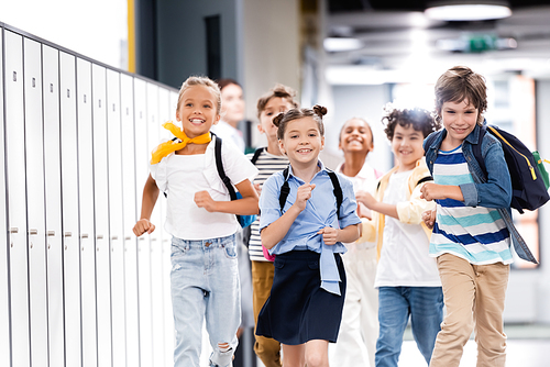 selective focus of excited multicultural classmates with backpacks running along school corridor