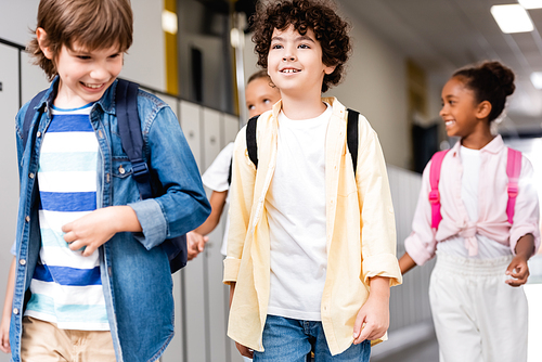 excited multicultural schoolchildren walking along school corridor