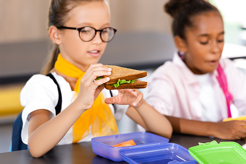 selective focus of schoolgirl in eyeglasses holding sandwich near african american classmate in school eatery