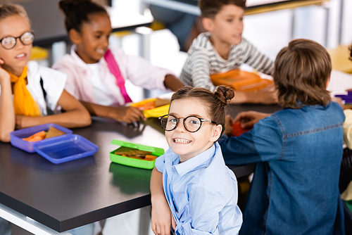 selective focus of excited schoolgirl in eyeglasses  in school canteen near multiethnic classmates