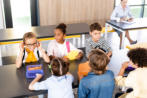 multiethnic classmates sitting in school canteen near lunch boxes and teacher on background