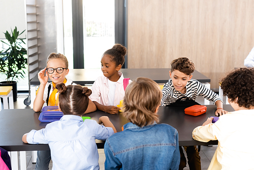 schoolgirl touching eyeglasses while sitting with multicultural classmates in school eatery