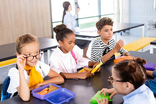 schoolgirls talking while sitting in school eatery with multicultural classmates and teacher on background