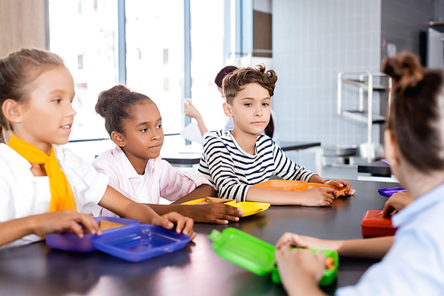 selective focus of multicultural pupils sitting in school canteen near lunch boxes