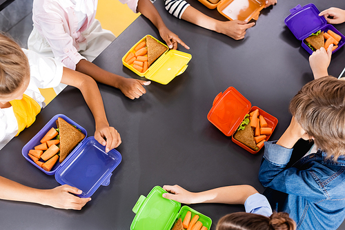 overhead view of multicultural classmates sitting in school eatery near lunch boxes with fresh carrots and sandwiches