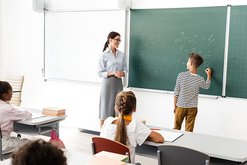 back view of schoolboy solving equations on chalkboard near teacher and multiethnic pupils