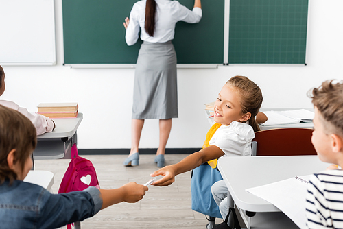 selective focus of pupil passing note to schoolgirl while teacher writing on chalkboard during lesson