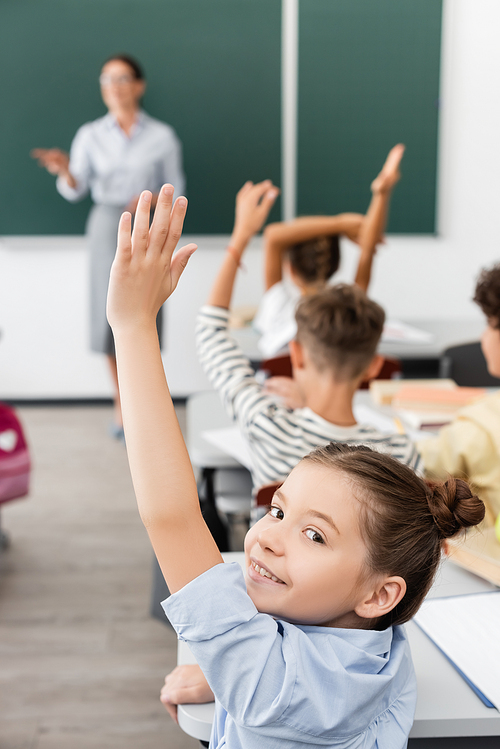 selective focus of schoolgirl with hand in air  near classmates and teacher on background