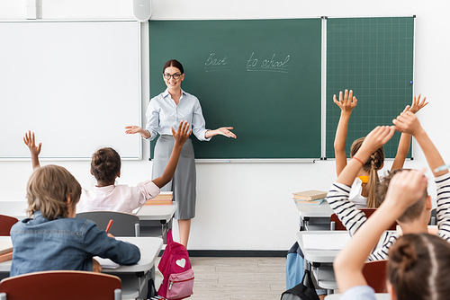 back view of multiethnic pupils with hands in air, and teacher standing with open arms near chalkboard with back to school lettering