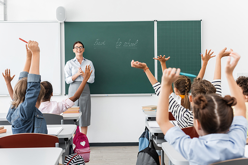 back view of multicultural pupils with hands in air, and teacher standing at chalkboard with back to school lettering
