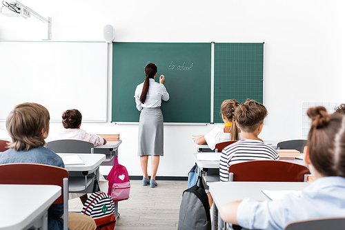 back view of teacher in formal wear writing on chalkboard near multiethnic pupils in classroom