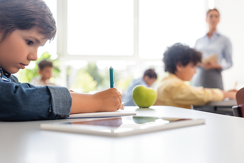 selective focus of schoolboy writing during lesson near apple and digital tablet