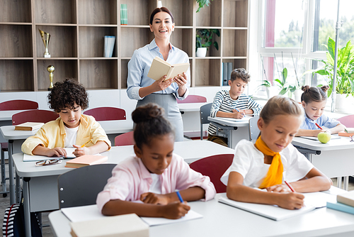 teacher holding book and  while multicultural pupils writing dictation in classroom