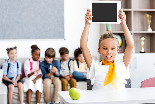 Selective focus of schoolgirl showing digital tablet near apple on desk and multiethnic classmates in classroom