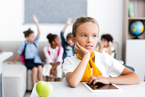 Selective focus of schoolgirl looking away near digital tablet and apple on desk in classroom