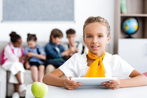 Selective focus of schoolgirl holding digital tablet near apple on desk and multiethnic friends in classroom