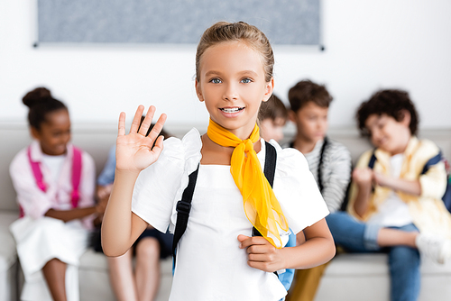 Selective focus of schoolgirl with backpack waving hand at camera with multicultural friends at background
