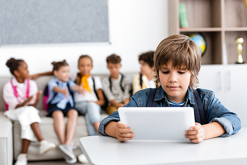 Selective focus of schoolboy holding digital tablet at desk with multicultural friends at background