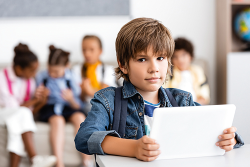 Selective focus of schoolboy holding digital tablet and  at desk in classroom