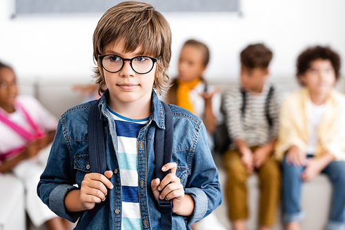 Selective focus of schoolboy in eyeglasses holding backpack in classroom