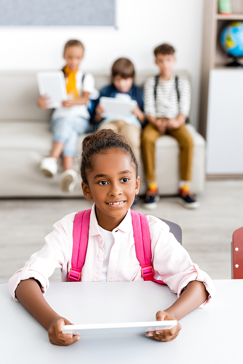 Selective focus of african american schoolgirl with backpack holding digital tablet at desk near friends in classroom