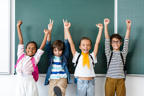 Selective focus of multicultural classmates showing peace and yes gestures near chalkboard in classroom