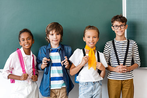 Multicultural schoolchildren with backpacks  near chalkboard