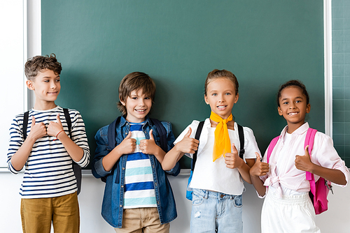 Multicultural schoolchildren showing thumbs up near chalkboard in school