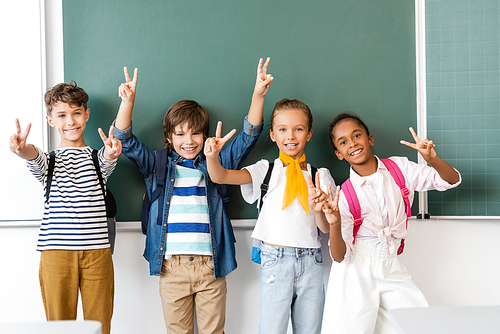 Selective focus of multiethnic schoolkids showing victory sign at camera near chalkboard in classroom