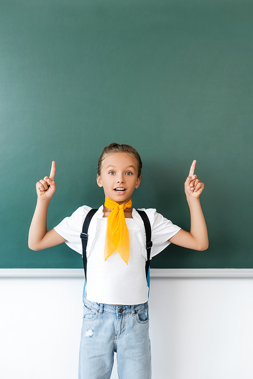 Excited schoolgirl with backpack pointing with fingers at chalkboard