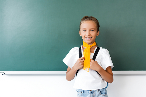 Schoolgirl holding backpack and  near green chalkboard