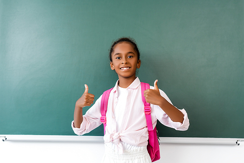 African american schoolgirl showing thumbs up near chalkboard in classroom