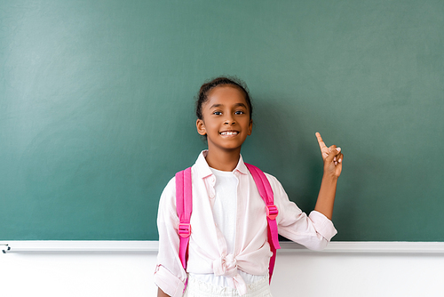African american schoolchild with backpack  while pointing with finger near chalkboard