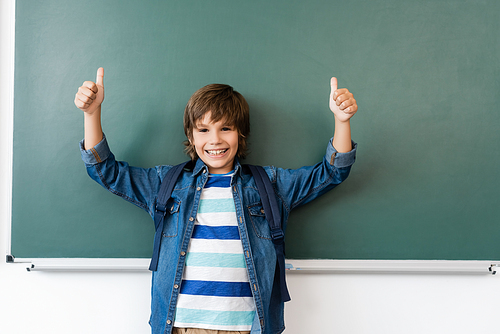 Schoolboy with backpack showing thumbs up near green chalkboard