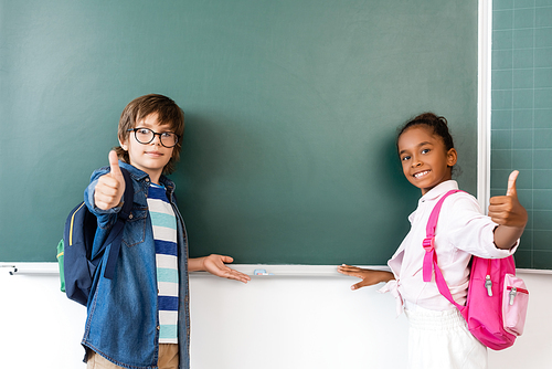 Multiethnic schoolchildren showing thumbs up at camera near chalkboard
