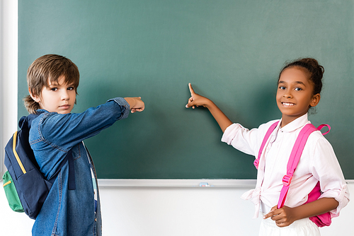 Multicultural schoolkids  while pointing at chalkboard in classroom
