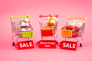 colorful presents in shopping trolleys with sale tags on pink, black friday concept