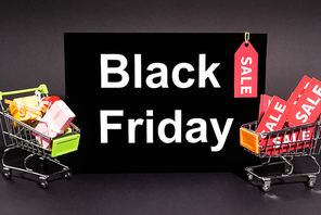 small presents in toy shopping carts near placard with black friday lettering and sale tag on dark background