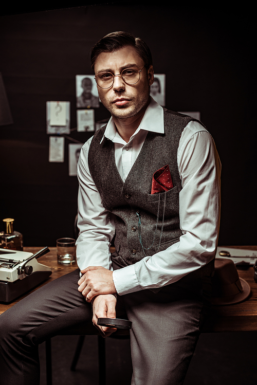 Detective in glasses sitting on table and holding magnifier in dark office