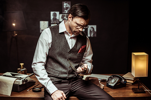 Concentrated detective in glasses sitting on wooden table and looking at pocket watch in office