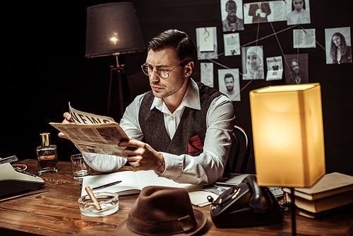 Concentrated detective in glasses reading newspaper in dark office