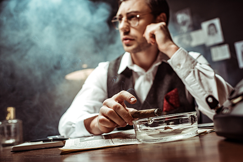 Selective focus of pensive detective in glasses holding cigar in office