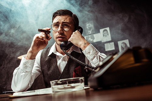 Detective in glasses holding cigar and talking on telephone in dark office