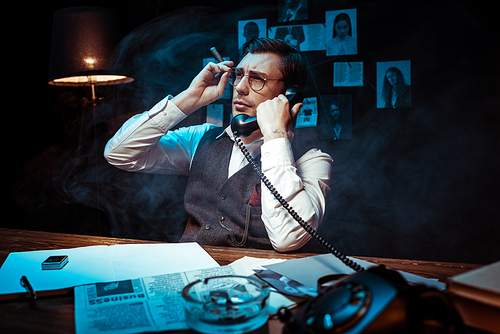 Detective in glasses holding cigar and talking on telephone in dark office