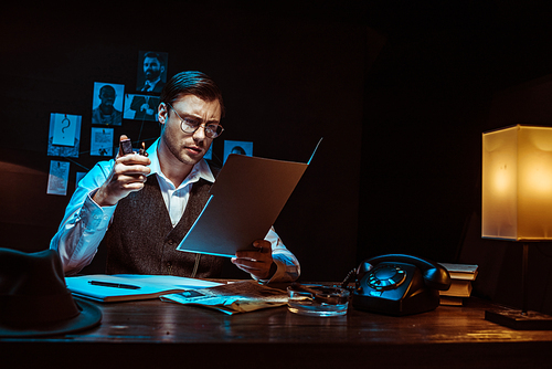 Concentrated detective in glasses reading dossier in dark office